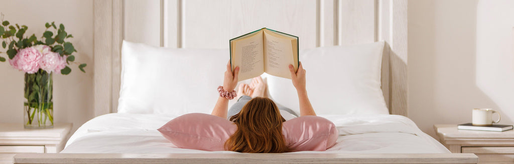 Woman laying on her back on a Rose Pink silk pillowcase reading a book on a bed.
