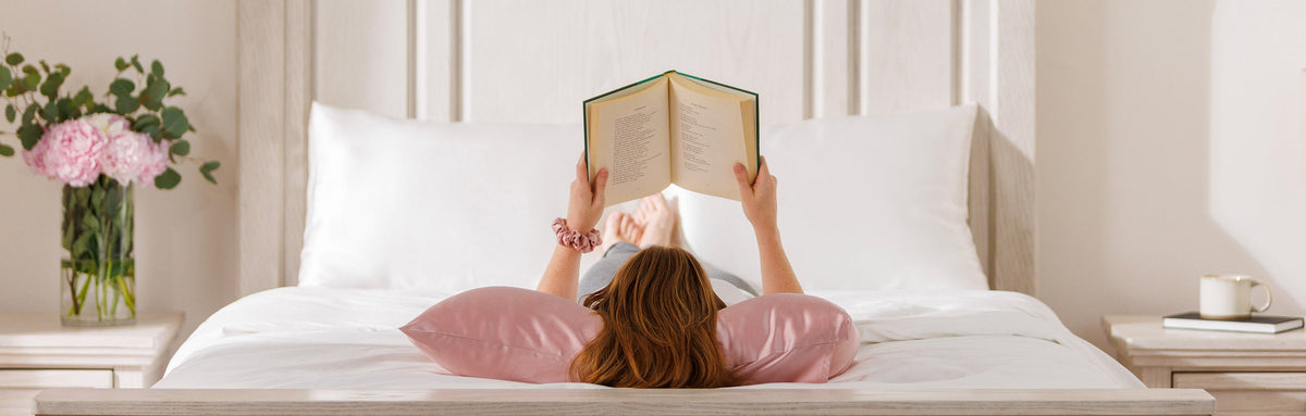 Woman laying on her back on a Rose Pink silk pillowcase reading a book on a bed.