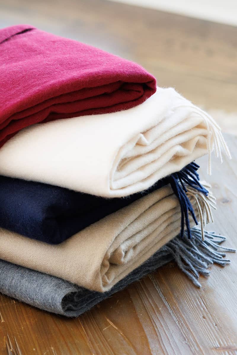 Stack of cashmere throws in cabernet, warm sand, navy, warm stone, and gray on wooden surface.
