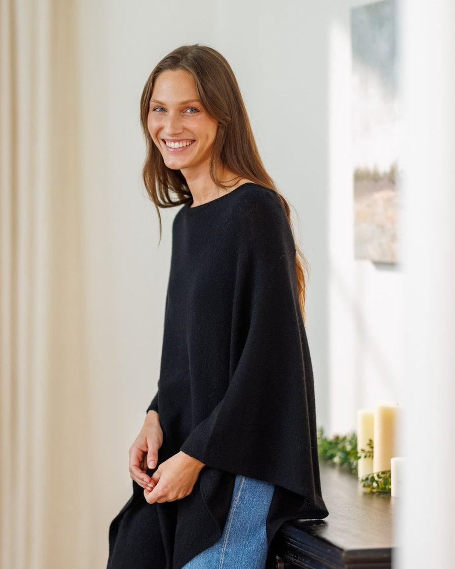 Woman in a black cashmere poncho smiling by a sunlit window