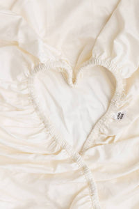 A close-up of a cream-colored fitted sheet with a heart-shaped elasticized cutout, showing the gathered fabric texture and a small tag that says "Long Side".