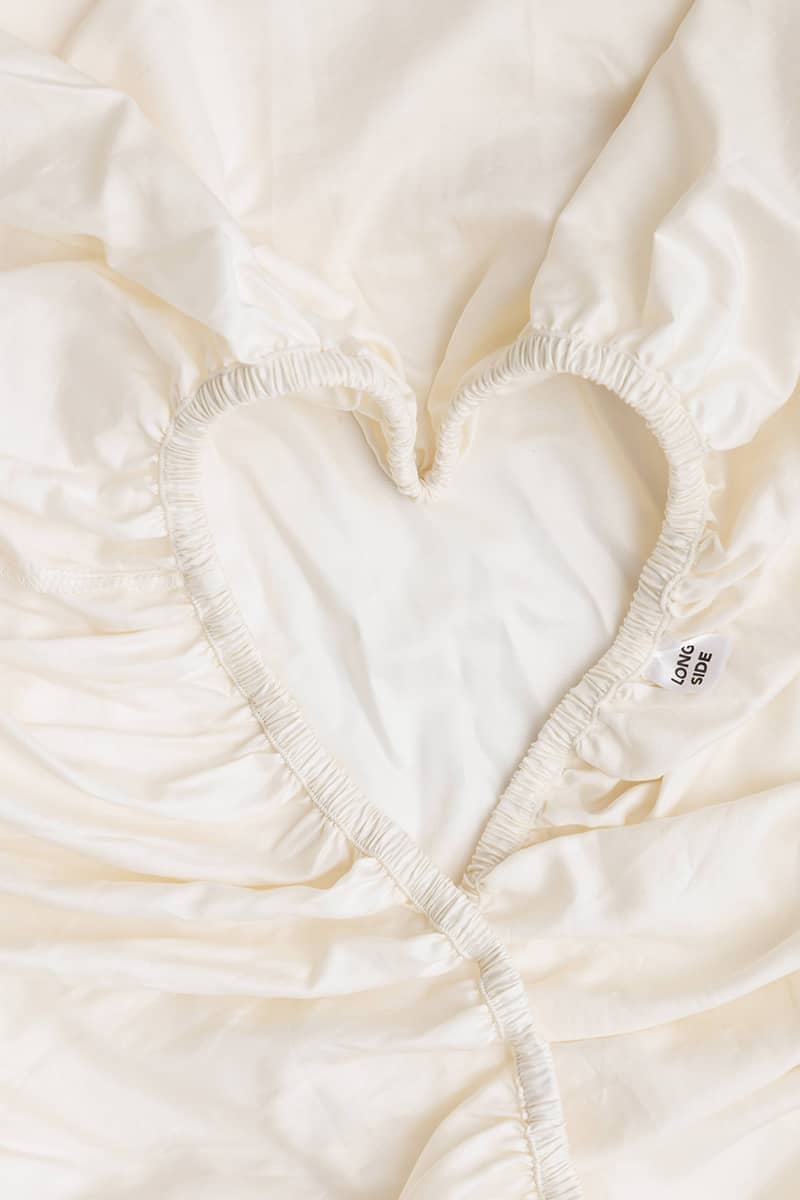 A close-up of a cream-colored fitted sheet with a heart-shaped elasticized cutout, showing the gathered fabric texture and a small tag that says "Long Side".