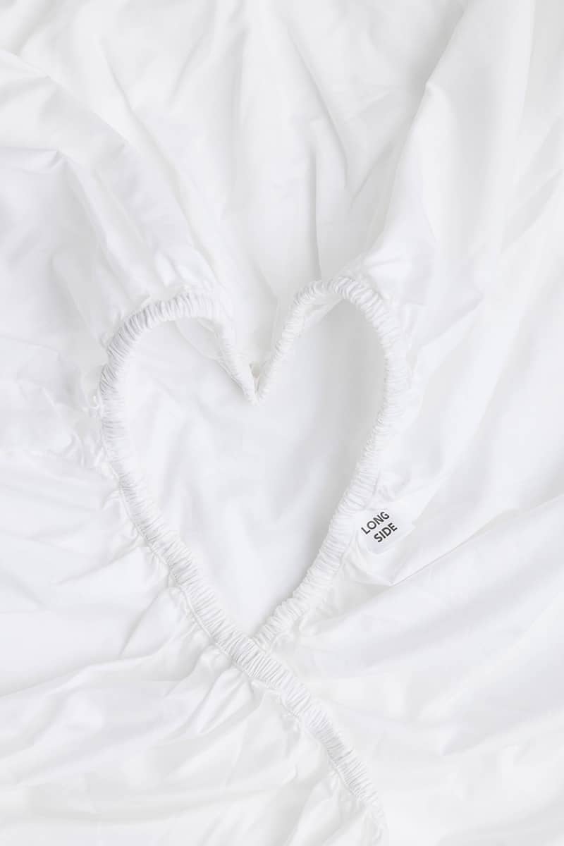 A close-up of a white fitted sheet with an elasticized band, featuring a heart-shape and a small tag that says "Long Side".