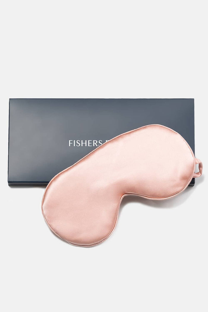 Blush pink silk sleep mask with adjustable strap next to navy Fishers sleepwear box.