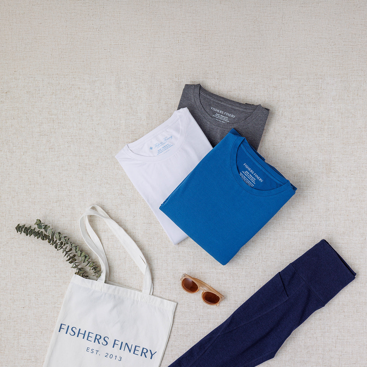 Folded shirts, a tote bag, and sunglasses on a textured surface with 'Fishers Finery' branding.