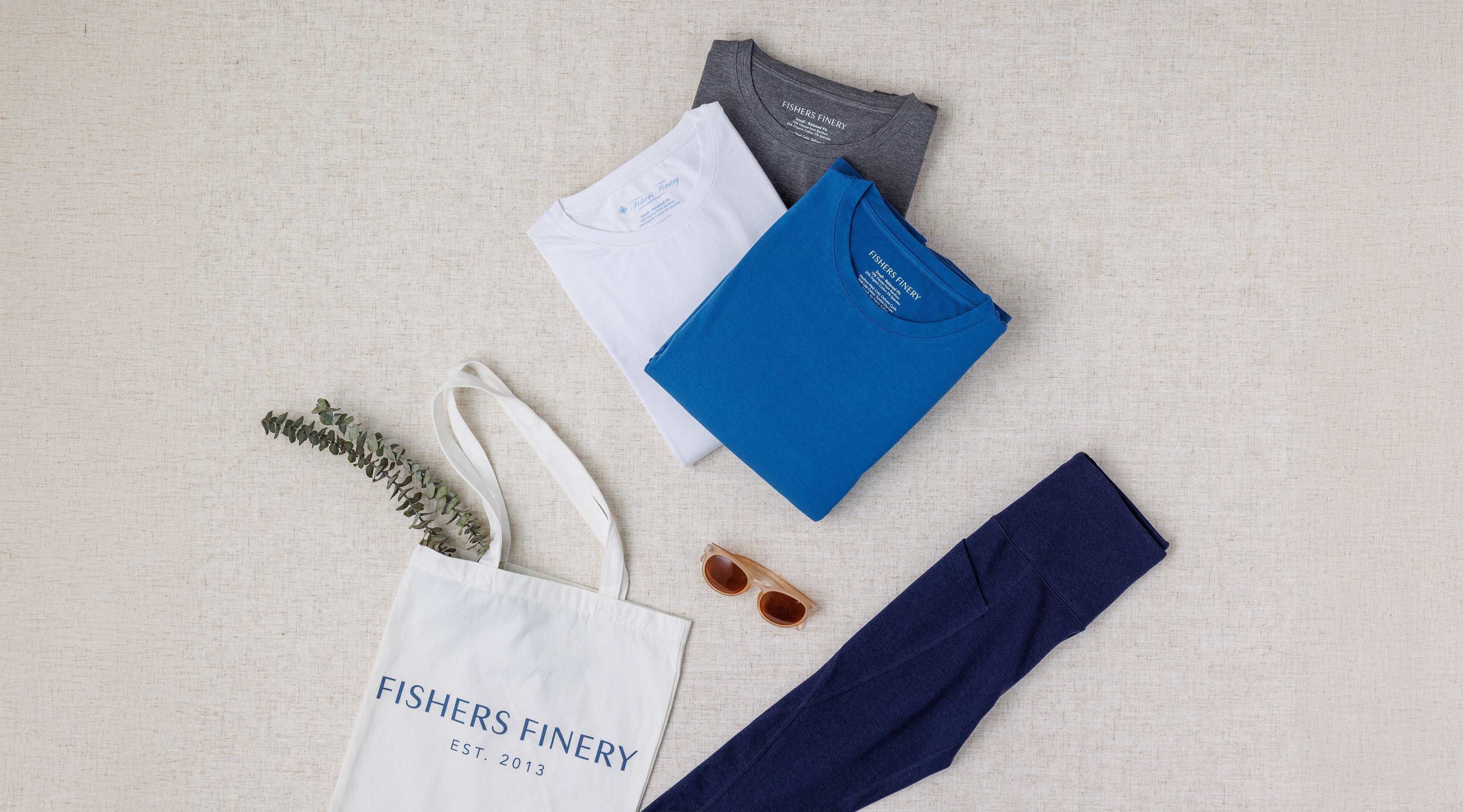 Folded shirts, a tote bag, and sunglasses on a light gray surface with 'Fishers Finery' branding.
