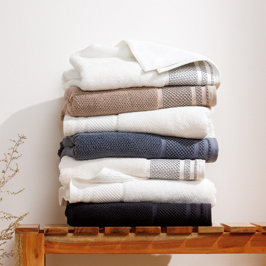 Stack of Rice Weave organic cotton towels in white, tan, charcoal and black on a wooden bench