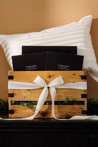 stripped white silk pillowcase in pillow in a wooden crate with black gift boxes. Crate wrapped in white silk bow with green leaves around box