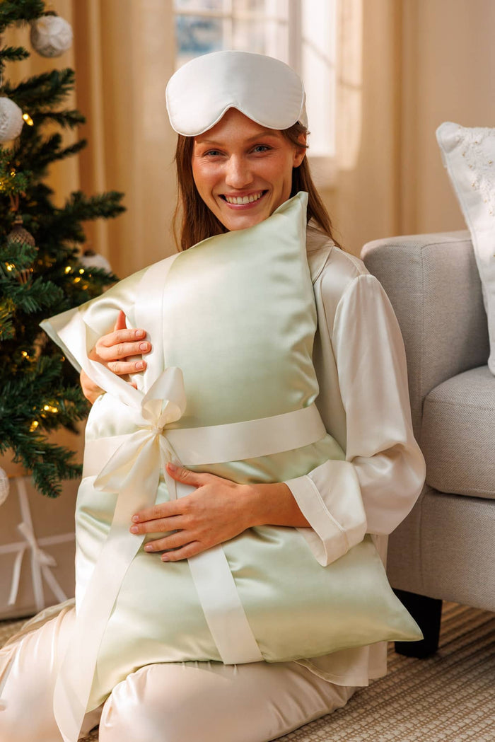 woman embracing a light green silk pillowcase wrapped in a white bow with a decorated tree behind her. She is wearing a white pj set and silk sleep mask