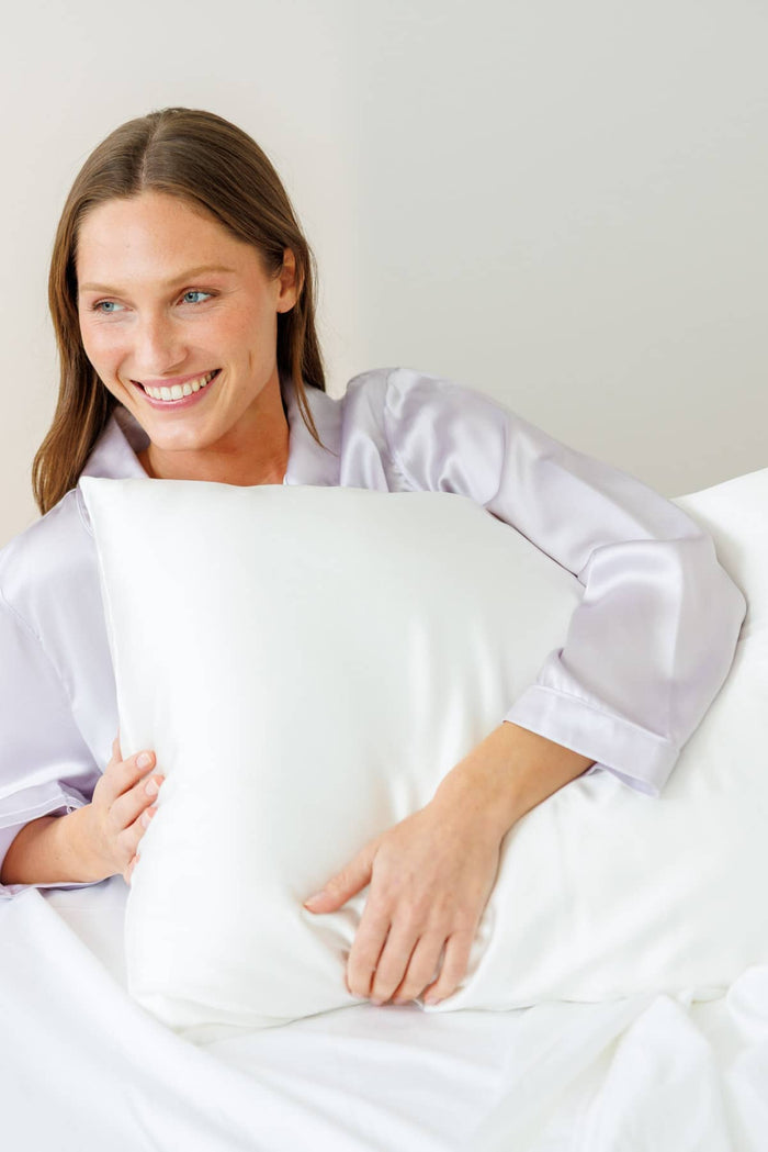 women laying in bed wearing purple silk pajamas while holding a white silk pillowcase