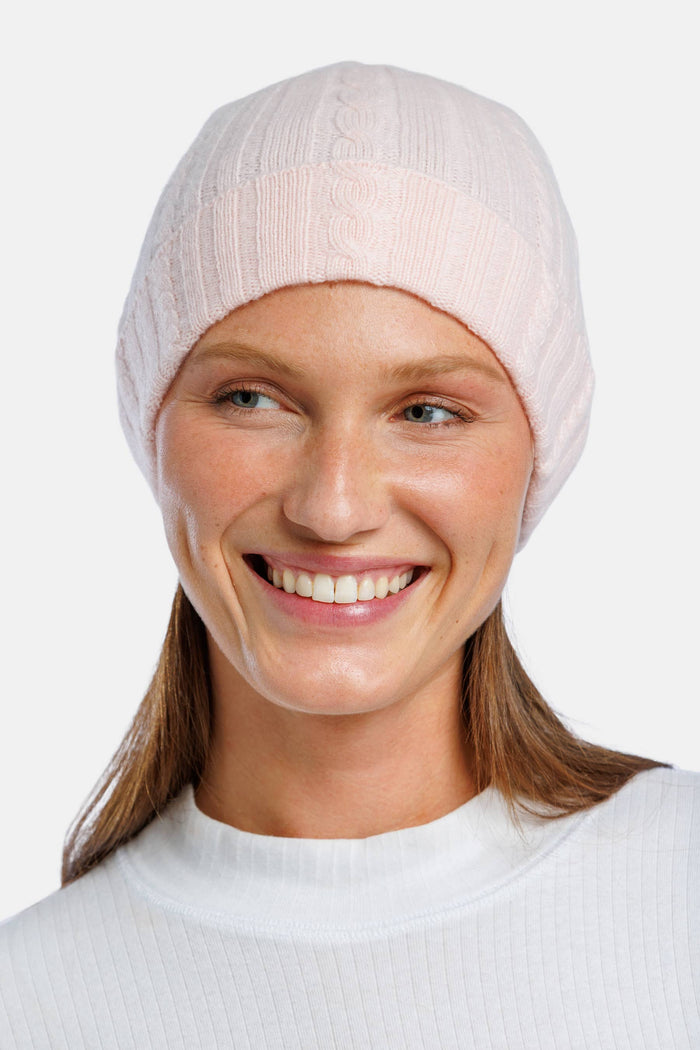 A smiling woman wearing a light pink cable knit beanie hat, shown from the chest up against a plain light gray background.