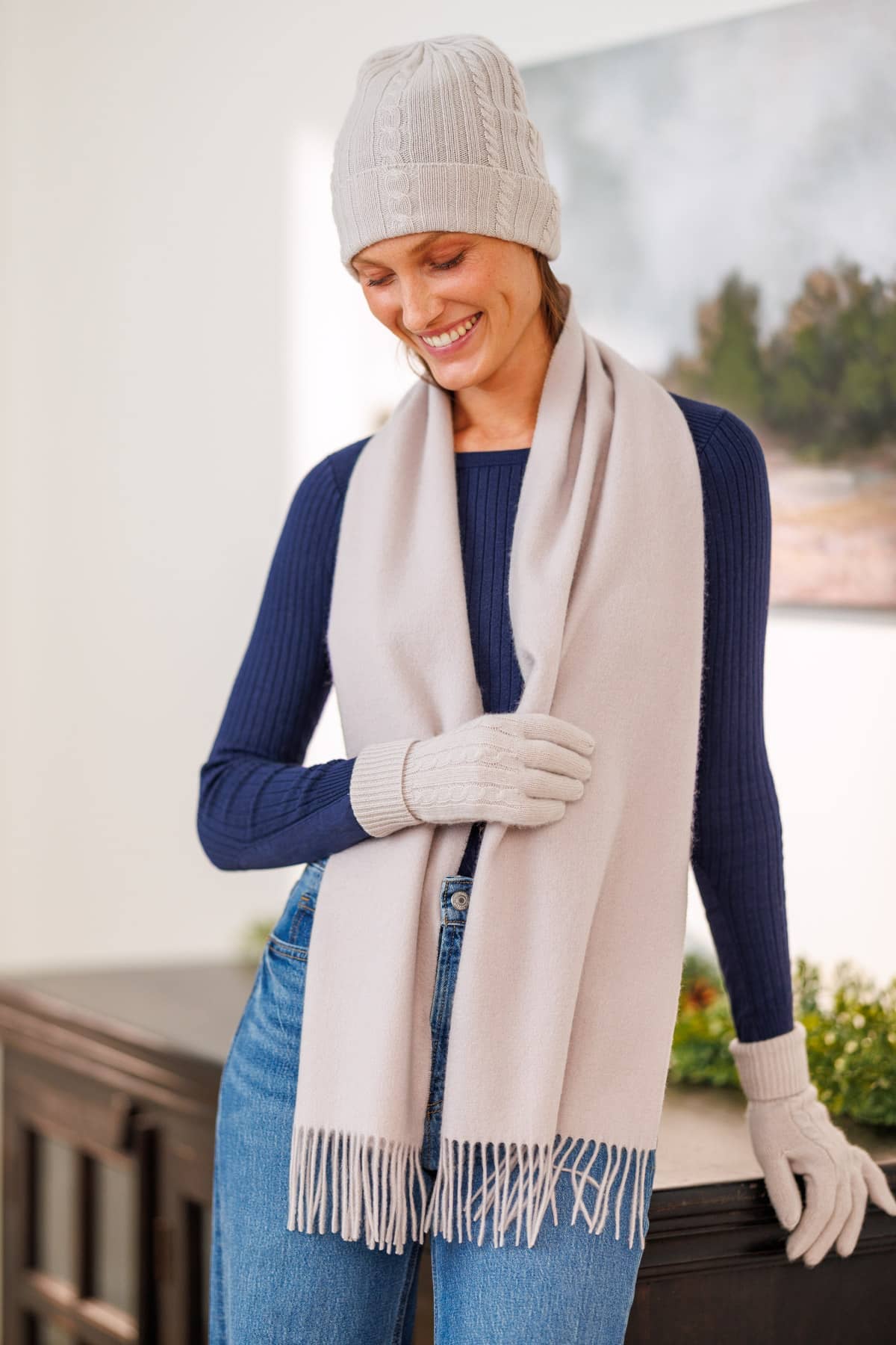 women wearing a light gray cashmere hat glove and scarf set over a dark blue top and jeans leaning on a black cabinet