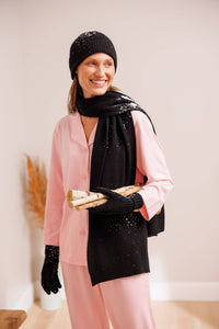 Woman wearing pink pajamas with black cashmere hat glove and scarf set carrying wood with snow flakes on the cashmere set.