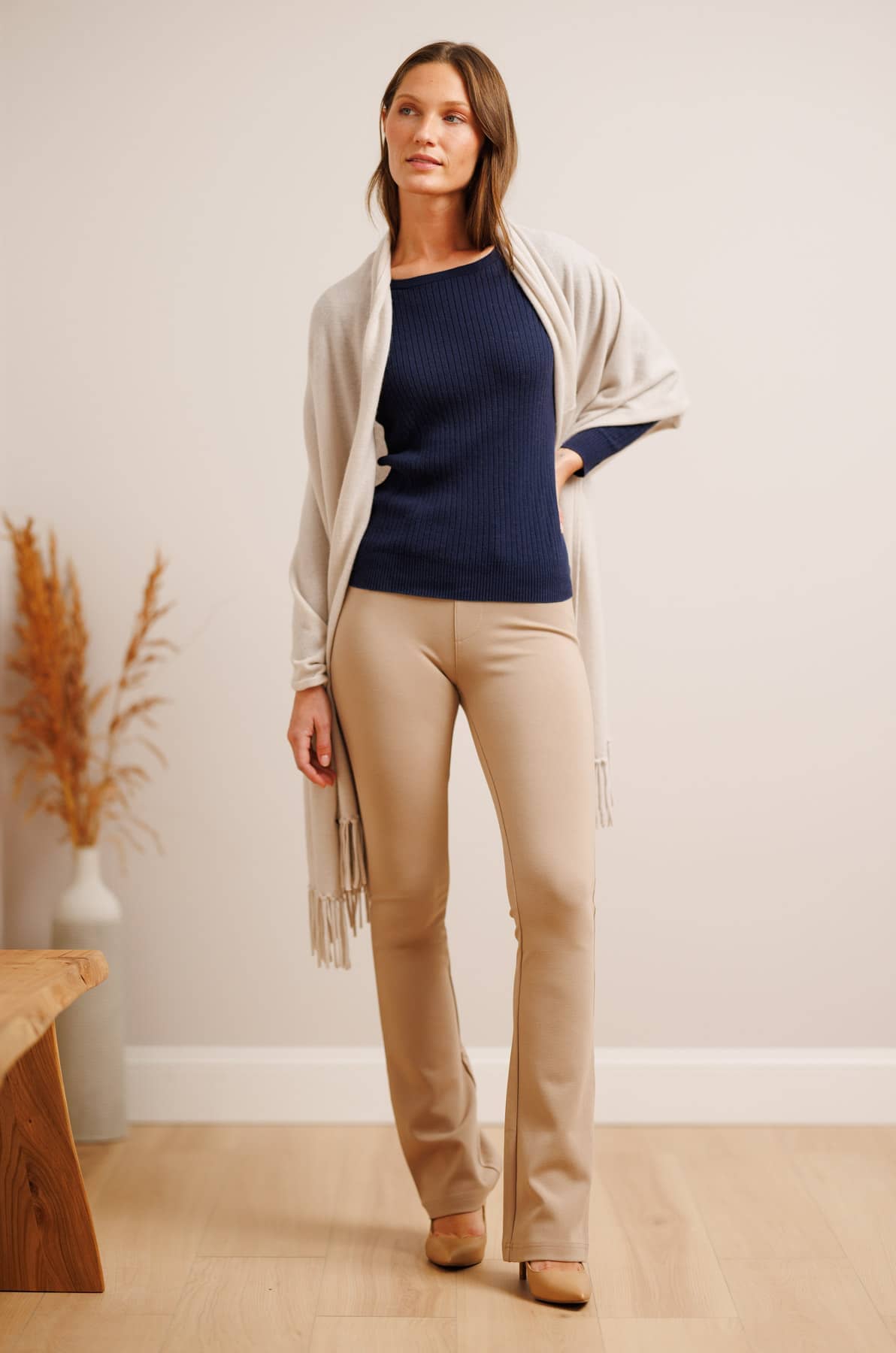 A woman models a coordinated outfit consisting of a navy ribbed knit top, beige straight-leg pants, and a light beige open-front cardigan with fringe, standing in a minimalist room with light wood flooring and a neutral wall.