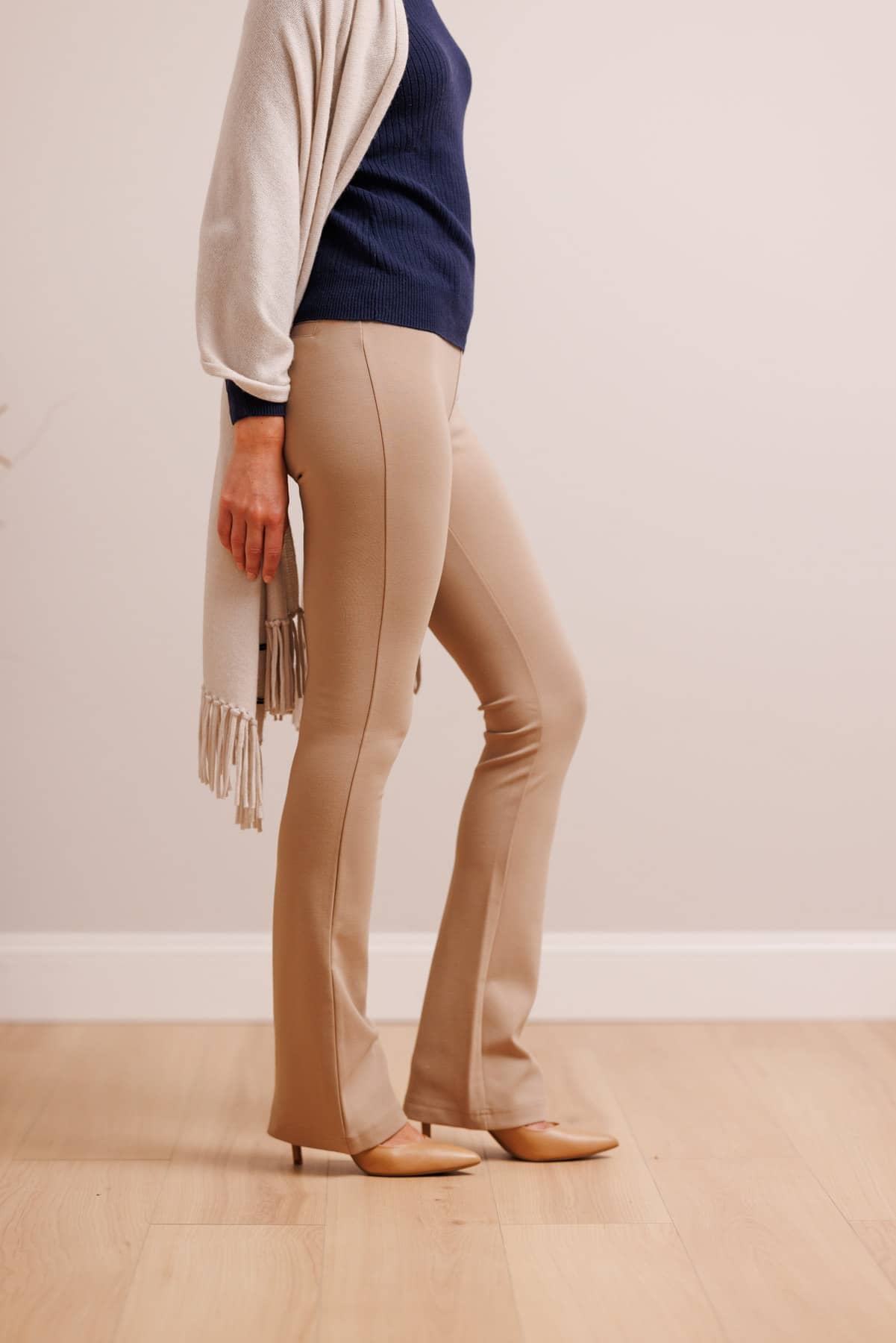 A model wearing beige flared pants, a navy blue top, a cream cardigan, and beige heels, standing in a room with light wood floors and a cream wall.