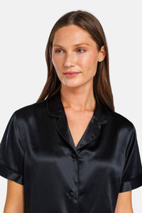 A woman wearing a black silk short-sleeve button-up shirt with a collar, shown from the chest up against a light gray studio background.