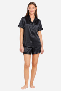 A woman modeling a black silk pajama set consisting of a short-sleeve button-front shirt and matching shorts, photographed against a plain light gray studio background.