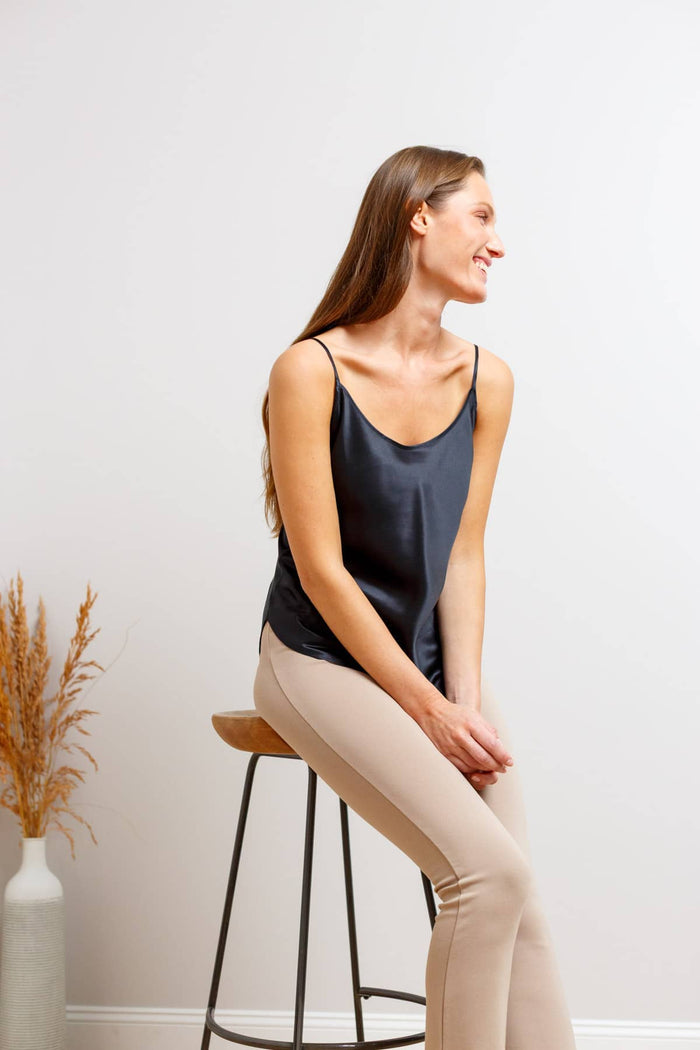 A woman with long brown hair sits on a wooden stool, wearing a dark black silk camisole and beige leggings, set against a plain light gray wall with a vase of dried grass on the left.