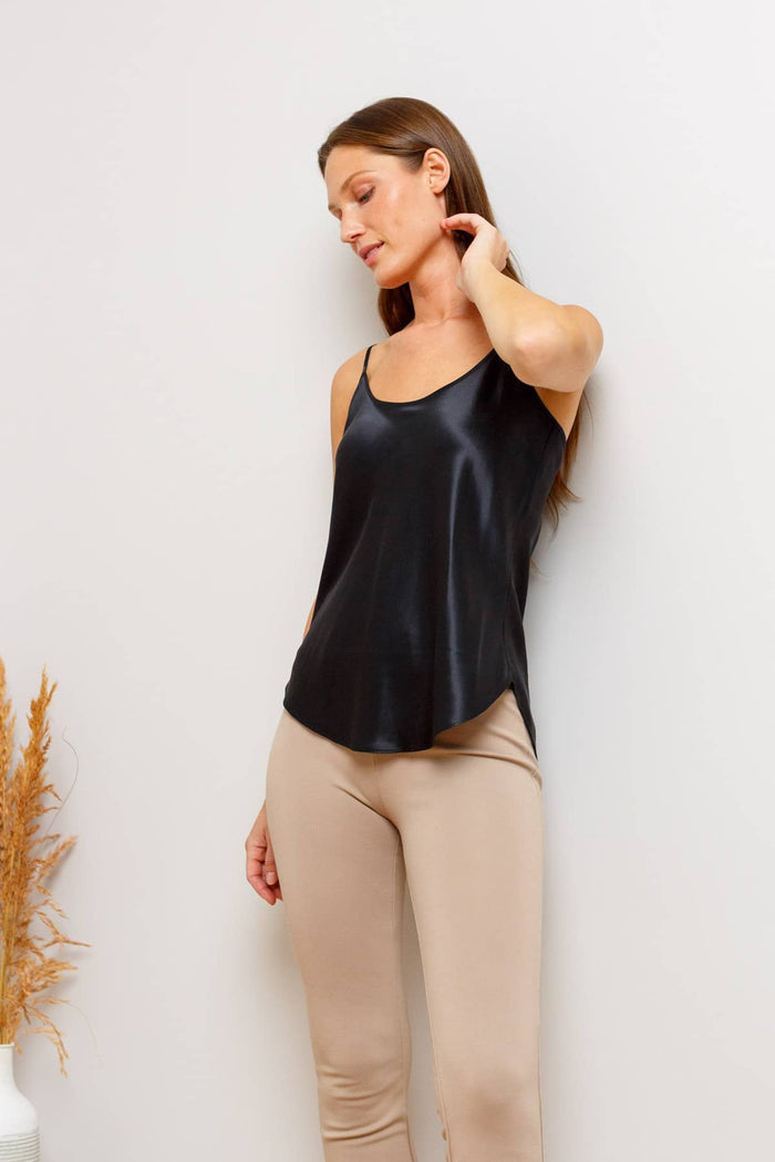 A woman wearing a black silk camisole tank top and beige leggings, posing against a plain white wall.