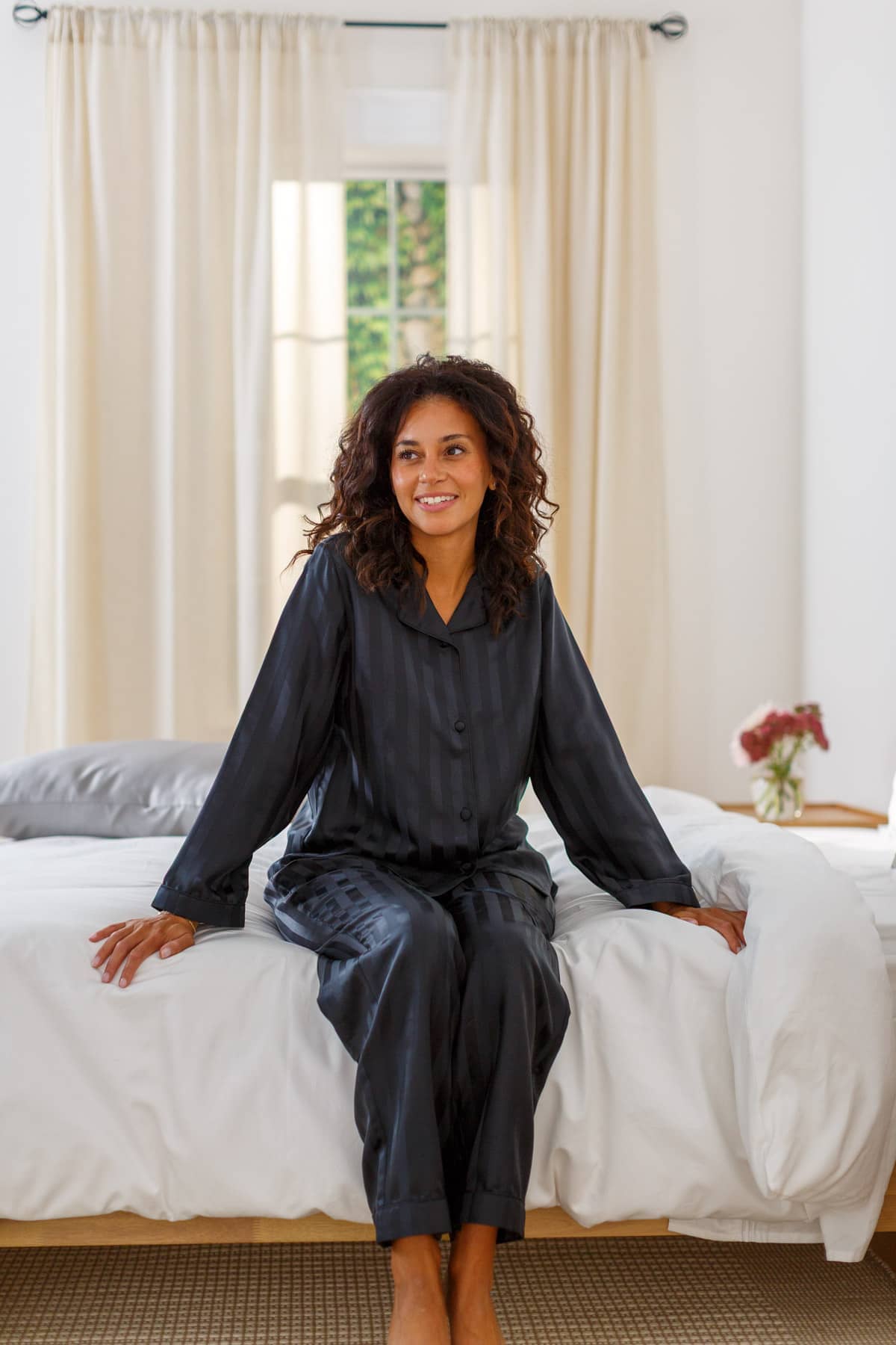 Woman wearing black striped mulberry silk full length pajama set while sitting on a bed