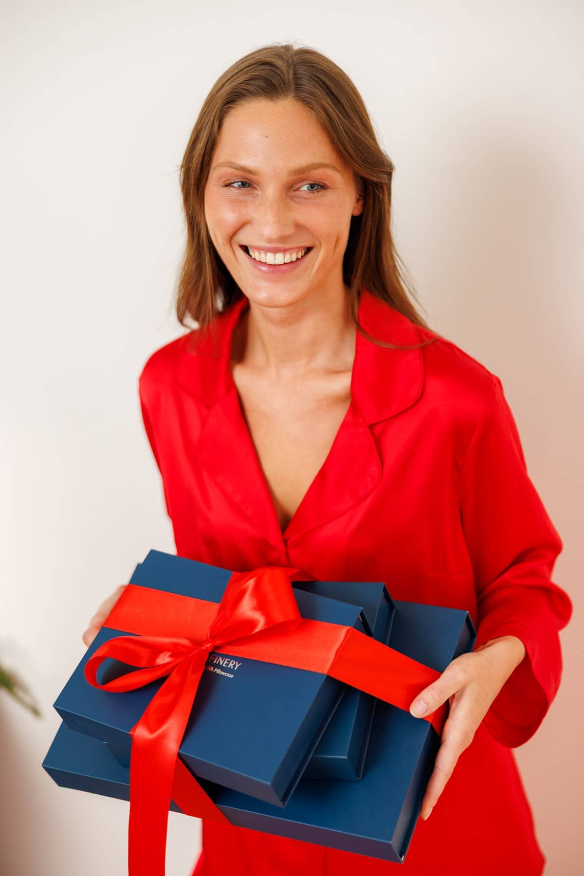 woman wearing a red silk pajama set while holding blue gift boxes wrapped in a red bow.